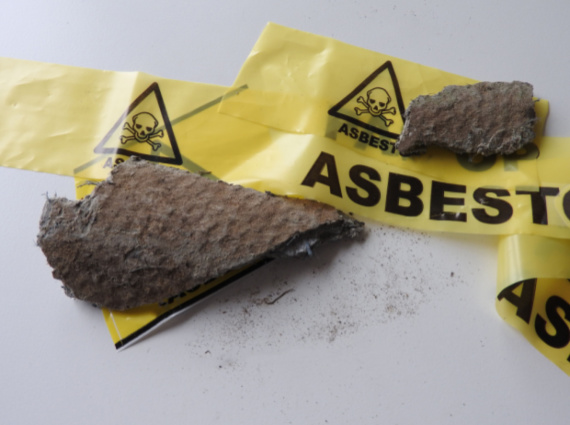 Pieces of asbestos material with visible fibers lying on a surface, surrounded by yellow caution tape labeled "asbestos" with hazard symbols, indicating an emergency restoration area.