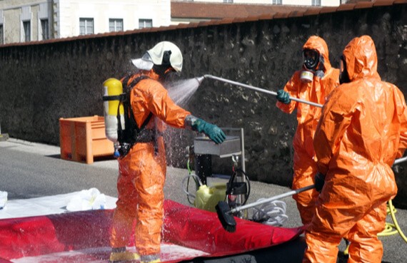Three workers in orange hazmat suits are conducting emergency restoration by decontaminating an area, spraying a substance with a hose, as one holds a monitoring device. A containment setup is visible in the