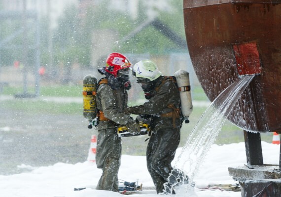 Two firefighters in full gear, wearing helmets with visible numbers, are using a hose together near a large metal structure, engaging in emergency restoration as water sprays around them amidst a snowy setting.