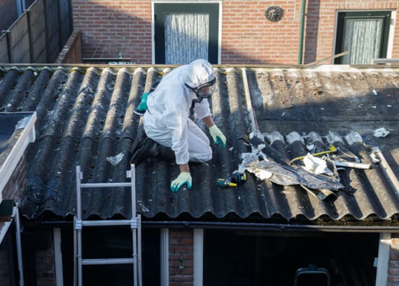 A worker in a protective suit and gloves performing emergency restoration by removing old roofing materials on a sloped, asbestos roof next to a metal ladder, with residential buildings in the background.