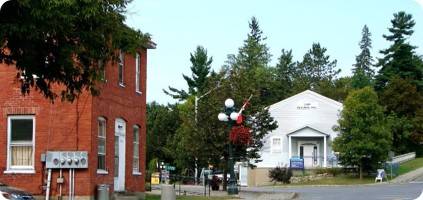 A small town street scene featuring a red brick emergency restoration building on the left, a white building on the right, and mailboxes in the foreground, surrounded by green trees.