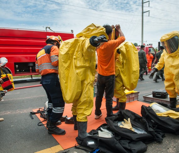 Emergency restoration responders in yellow hazmat suits prepare near a red fire truck, with some adjusting their gear on a road strewn with equipment.