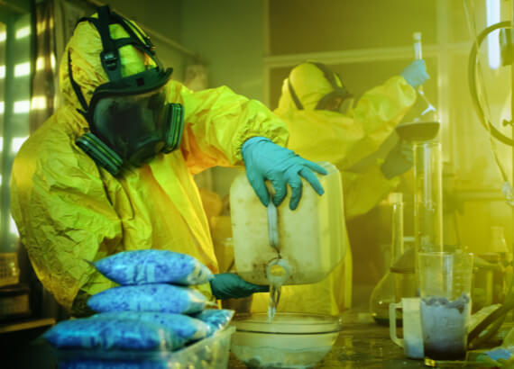 A person in a yellow hazmat suit and gas mask handling chemicals in an emergency restoration lab, pouring liquid from a container, surrounded by equipment and substances.