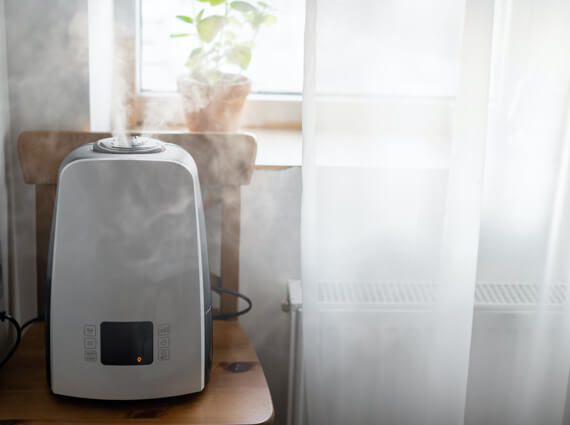 An ultrasonic humidifier operates on a wooden table by a sunny window with sheer curtains, aiding in emergency restoration. Visible mist emanates from the top, enhancing indoor air moisture.