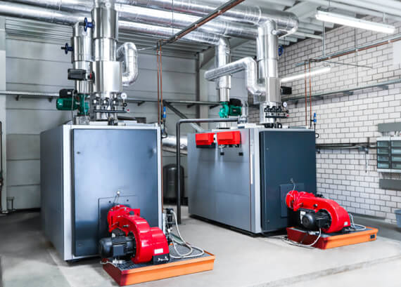 Industrial boiler room with two large metal boilers, red pumps, and an array of silver pipes and valves, within a clean, well-organized emergency restoration space with white tiled walls.