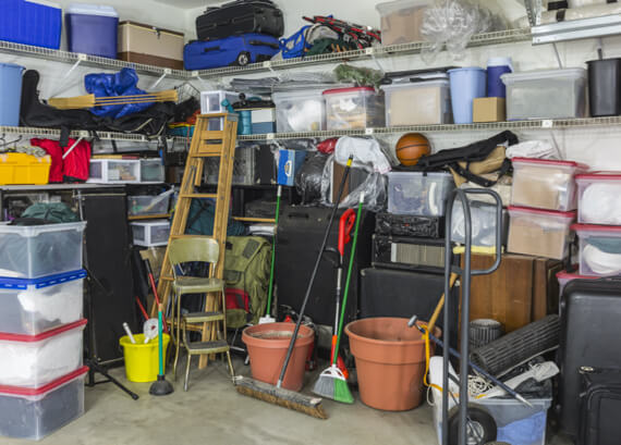 A cluttered garage with shelves filled with emergency restoration equipment, suitcases, and various household items like a ladder, brooms, and sports equipment.