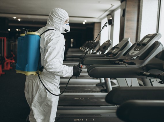A person in protective gear with a mask and backpack sprayer performs emergency restoration by disinfecting treadmills in a gym to ensure cleanliness and safety.