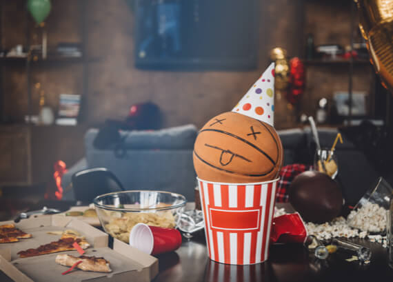 A basketball with a drawn face and party hat sits in a striped red and white emergency restoration bucket on a table littered with pizza slices, popcorn, and party debris, with a blurred festive background.