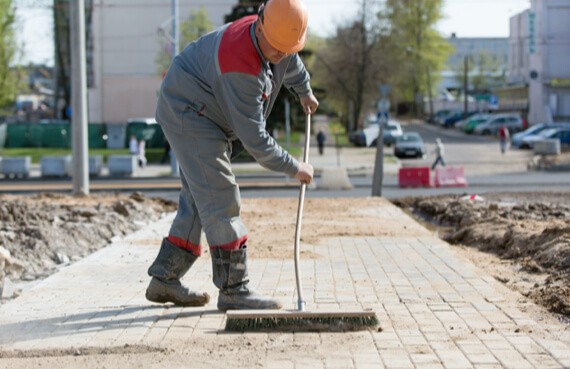 A construction worker in a gray uniform and orange helmet performs emergency restoration, sweeping sand between paving stones on a new sidewalk in an urban setting.