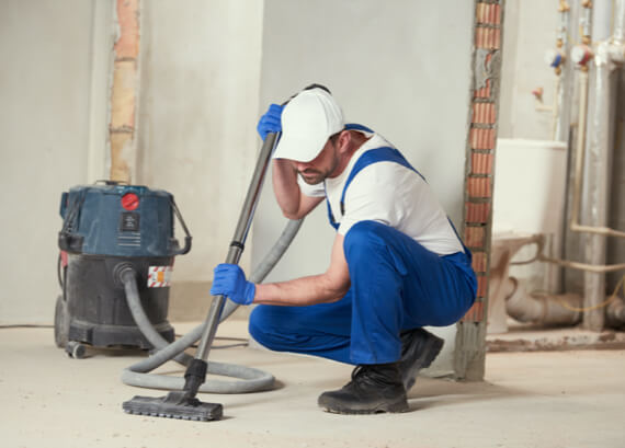 A construction worker in a white and blue uniform uses an industrial vacuum cleaner on a dusty floor in a partially renovated room during an emergency restoration.