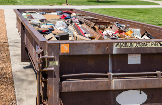 A large, full dumpster containing assorted discarded items like furniture and scrap metal, parked on a concrete surface beside a grassy area, ready for emergency restoration.