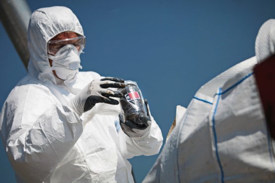 Two individuals in protective suits and goggles handling a potentially hazardous material in a sealed container during an emergency restoration, with a clear blue sky in the background.