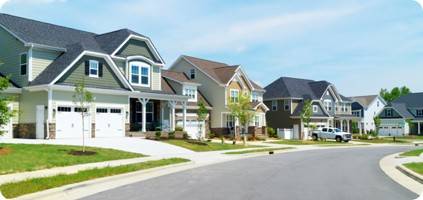 A row of new, modern suburban houses along a curving street on a sunny day, with well-manicured lawns and emergency restoration signs, under a clear blue sky above.