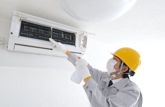 An emergency restoration technician in a helmet and mask services an air conditioner unit, inspecting it with a flashlight in hand.