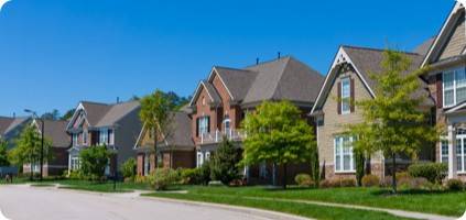 A row of upscale suburban homes with varied brick and siding facades under clear blue skies, each undergoing emergency restoration, with neatly manicured lawns and mature trees.