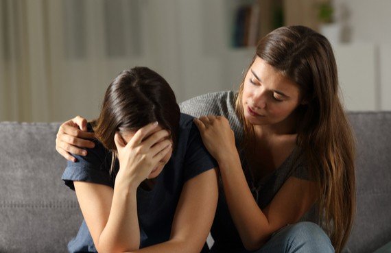 A woman comforts another who is visibly upset, holding her head in her hands, in a soothing embrace on a couch in an emergency restoration home setting.