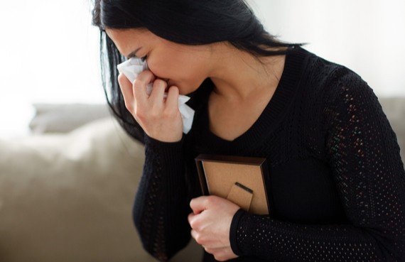 A woman in a black sweater is visibly upset, holding a tissue to her face and clutching a book or journal to her chest, sitting on a couch after an emergency restoration incident.