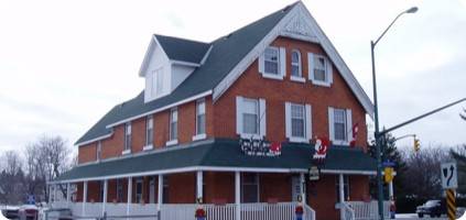 A large two-story brick building with white trim and a wrap-around porch, located at a street corner under a cloudy sky. The building displays several flags, an "Emergency Restoration" banner, and a