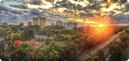 Panoramic cityscape at sunset with sunlight streaming through clouds, highlighting an emergency restoration road flanked by trees and high-rise buildings in the distance.